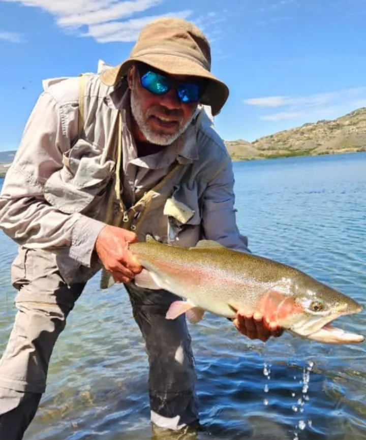 Pescador con sombrero y anteojos sosteniendo una trucha arcoíris en la orilla de un lago