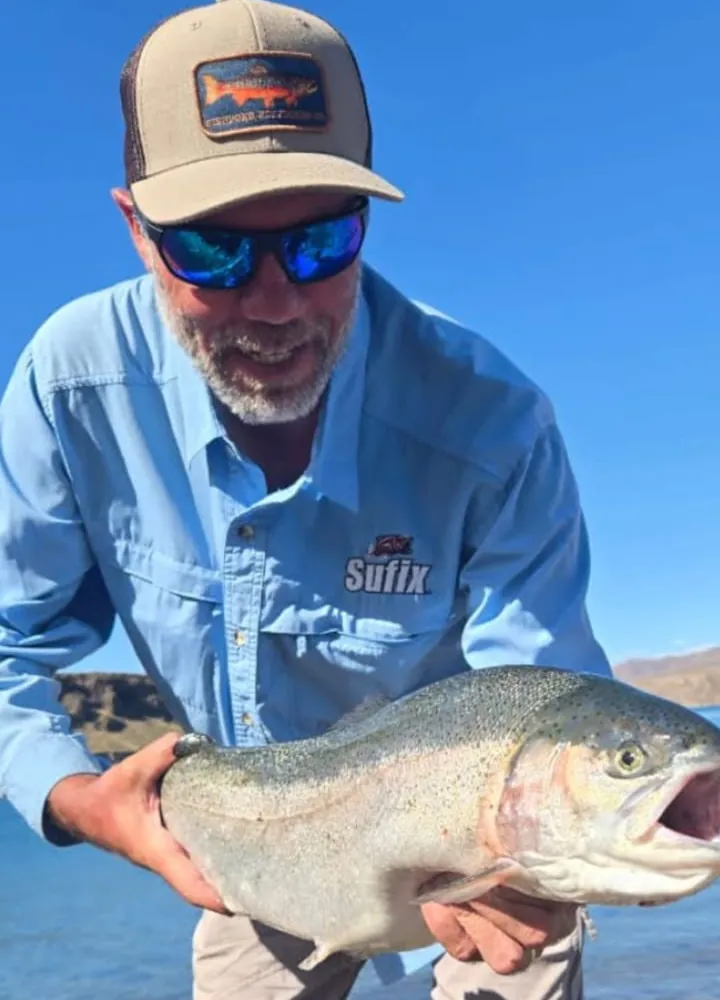 Pescador con gorra sosteniendo una trucha arcoíris grande junto al agua