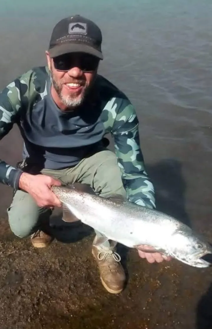 Pescador agachado en la costa sosteniendo una trucha plateada