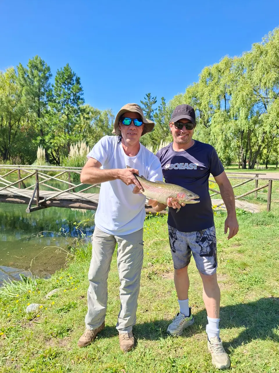 Dos pescadores sonrientes sosteniendo una trucha arcoíris junto a una laguna con un puente de madera