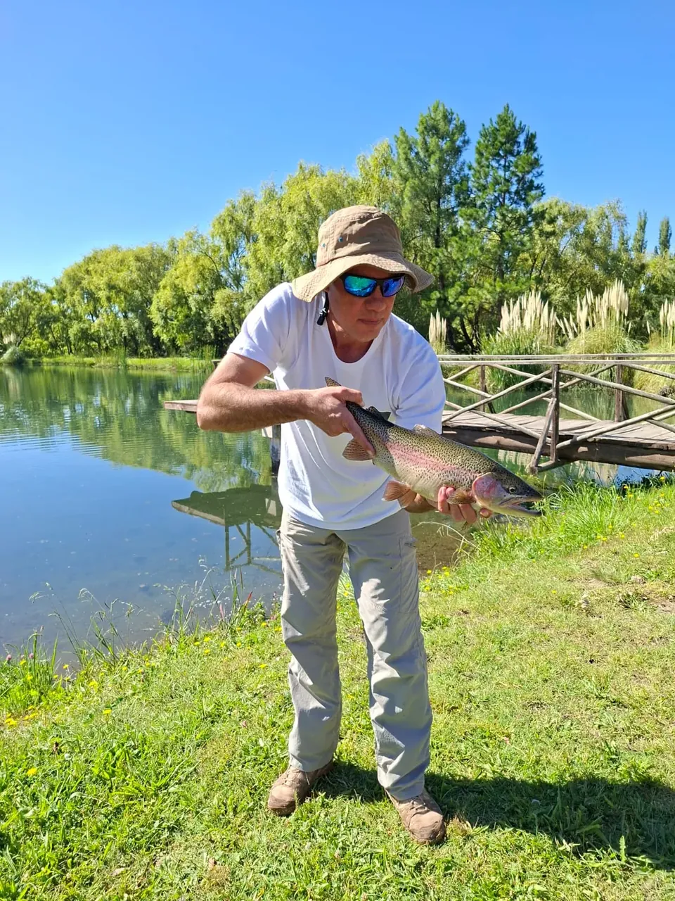 Pescador parado en el pasto junto a una laguna, sosteniendo una trucha arcoíris