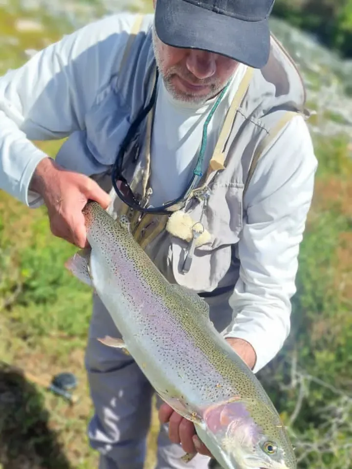 Pescador con gorra sosteniendo una trucha arcoíris grande entre los pastos de la orilla