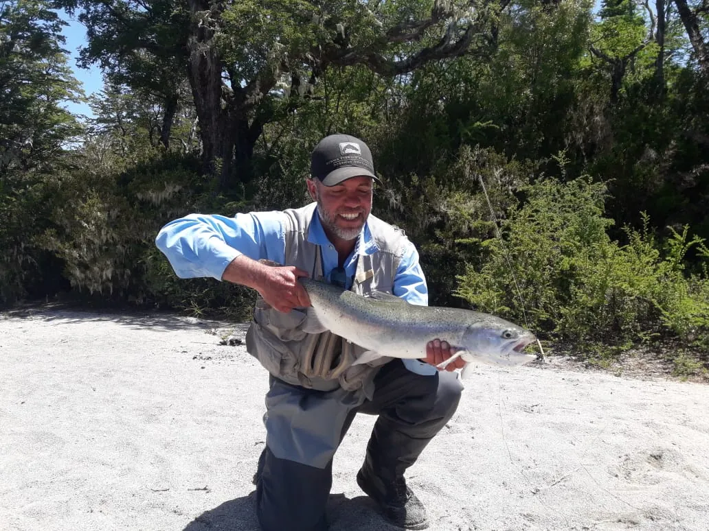 Pescador arrodillado en una playa de arena sosteniendo una trucha plateada