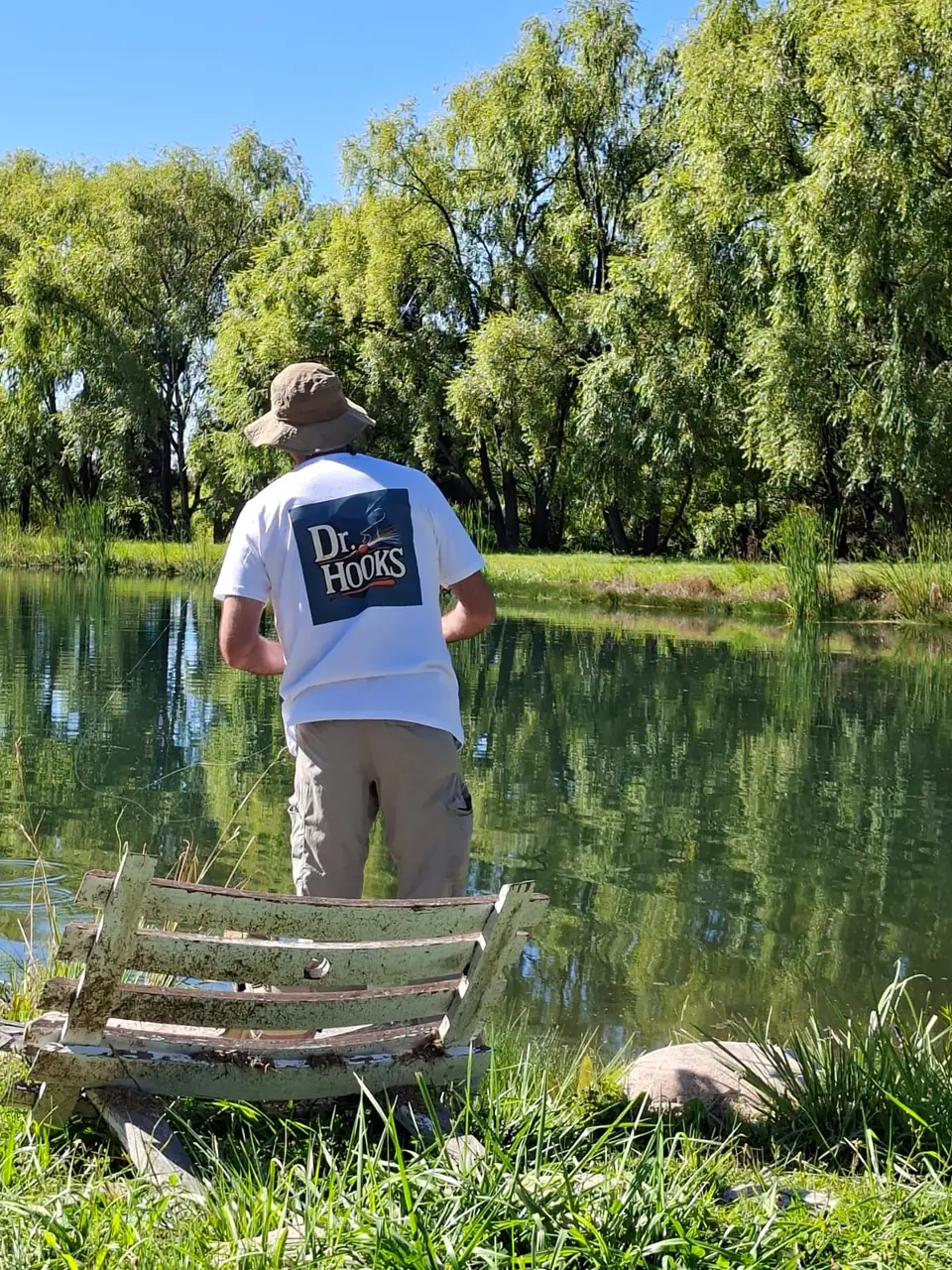 Pescador de espaldas con la remera de Dr. Hooks, pescando con mosca en una laguna rodeada de sauces